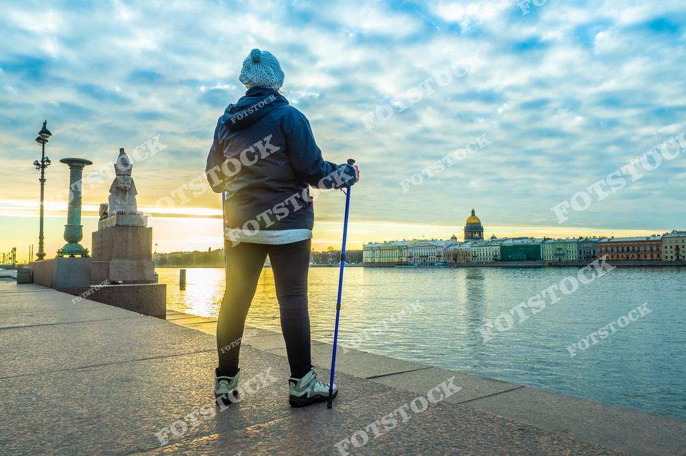 На фотографии изображена женщина, совершающая прогулку по Университетской набережной в Санкт-Петербурге. На заднем плане виден величественный Исаакиевский собор, один из символов города. По левую руку стоят знаменитые сфинксы, привезенные из Египта и установленные у Академии художеств. Раннее утро, отражения в реке Нева и светлое небо создают атмосферу спокойствия и умиротворения.